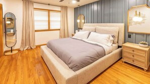 Un dormitorio con cama, suelo de madera, una mesita de noche, un espejo y una ventana con cortinas.