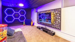 Habitación con televisor de pantalla plana grande, pared de luces hexagonales, sillón de peluche y dispensador de aperitivos.