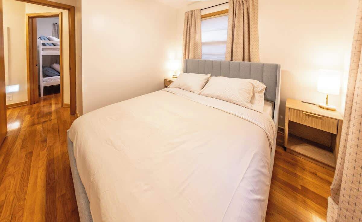 A neatly made bed with a grey headboard, a bedside table with a lamp, and a wooden floor.