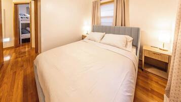 A neatly made bed with a grey headboard, a bedside table with a lamp, and a wooden floor.