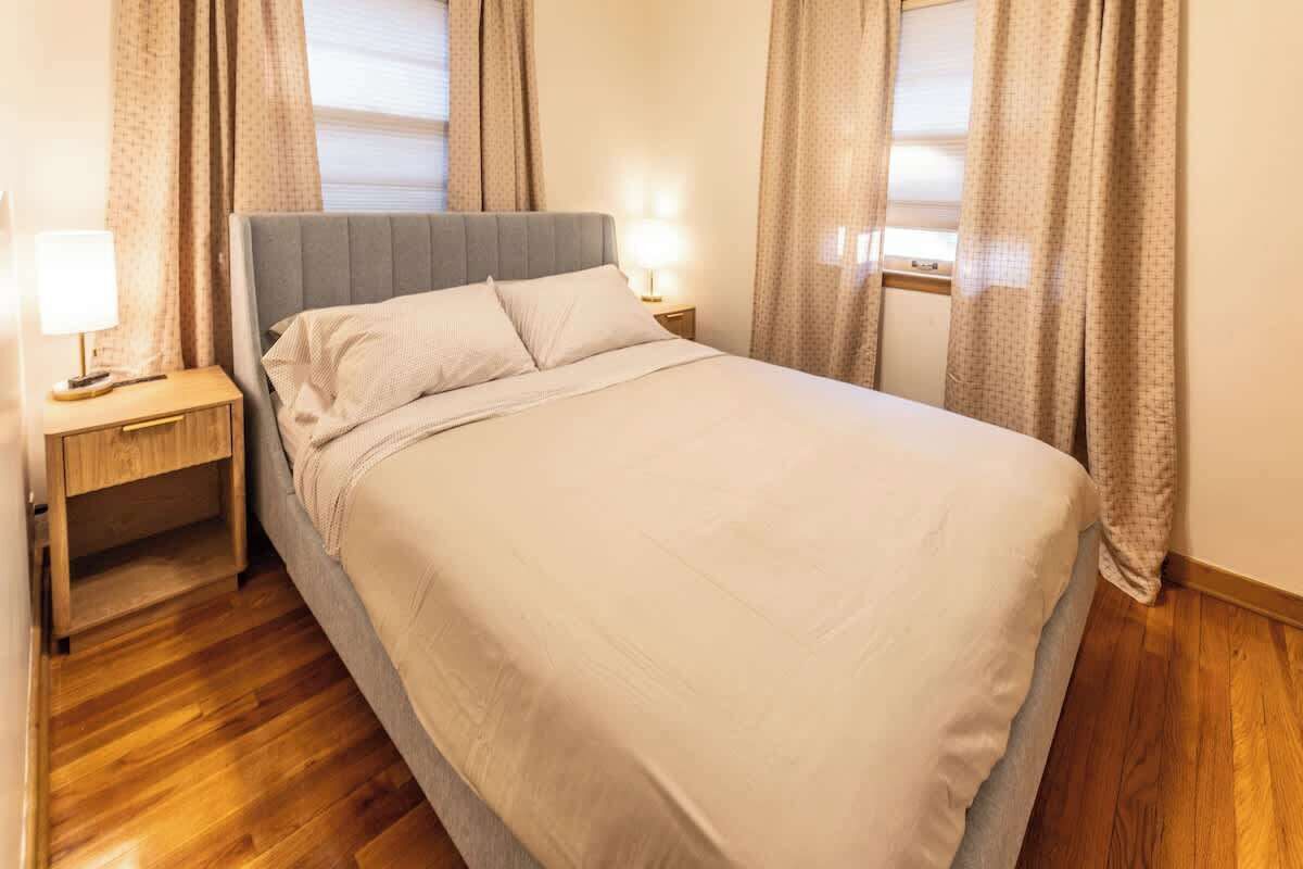 Een net opgemaakt bed met een grijs hoofdbord, een nachtkastje met een lamp en gordijnen voor de ramen.