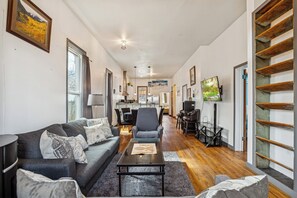 Una sala de estar moderna con un sofá, sillones, una mesa de centro y un televisor. Se observa un comedor con sillas y una mesa al fondo.