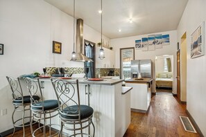 Una cocina moderna con una isla central, taburetes de bar, lámparas colgantes y vistas a un dormitorio.