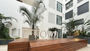 Un área de recepción moderna con un banco de madera, palmeras y un edificio con amplios ventanales al fondo.