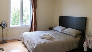 A bedroom with a bed, a bedside table, a nightstand, a standing fan, and a window with curtains.