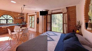 A bedroom with a large bed, a desk, and a chair. There is a window with curtains, a brick wall, and a television mounted on the wall. - Fika Cabin - Boscata (petfriendly) (El Oasis Valsequillo)
