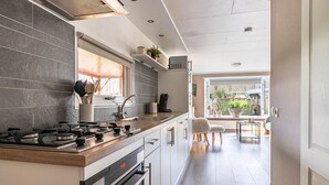 Eine moderne Küche mit Gasherd, Backofen und einem Fenster mit Jalousien. Ein Spülbecken mit Wasserhahn ist vorhanden, sowie eine Arbeitsfläche mit verschiedenen Gegenständen. Im Hintergrund sind ein Stuhl und ein kleiner Tisch zu sehen.