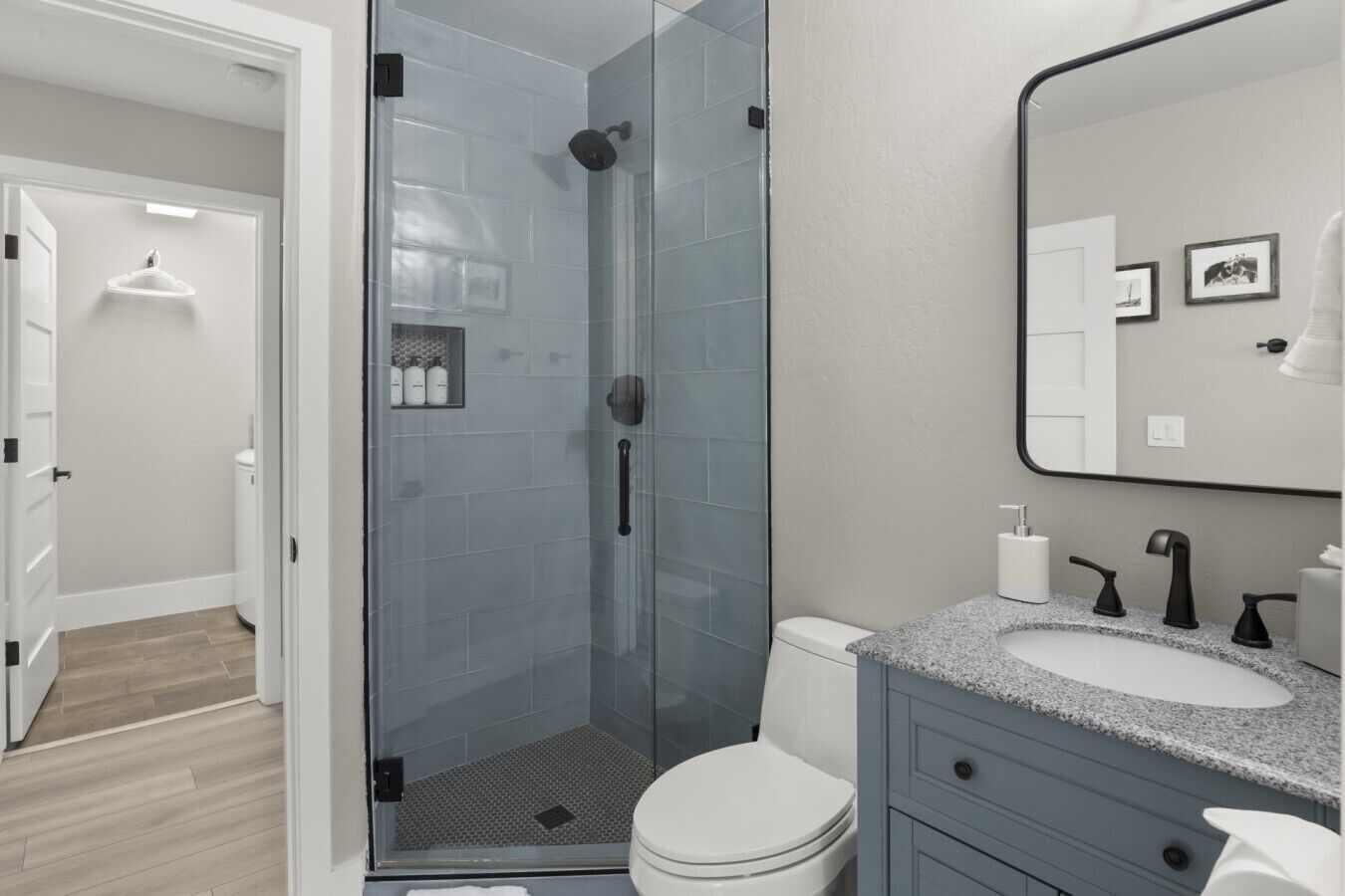 Une salle de bain moderne avec une douche à parois de verre, des toilettes blanches, un lavabo sur un plan de travail en granit et un miroir orné de cadres avec des photos.