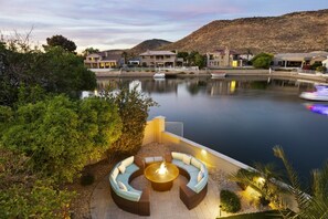 A lakeside property with a fire pit seating area, a boat dock, and multiple houses with a hilly backdrop. - Waterfront Pool, Dock & Theater | Paradise Palms (Glendale)