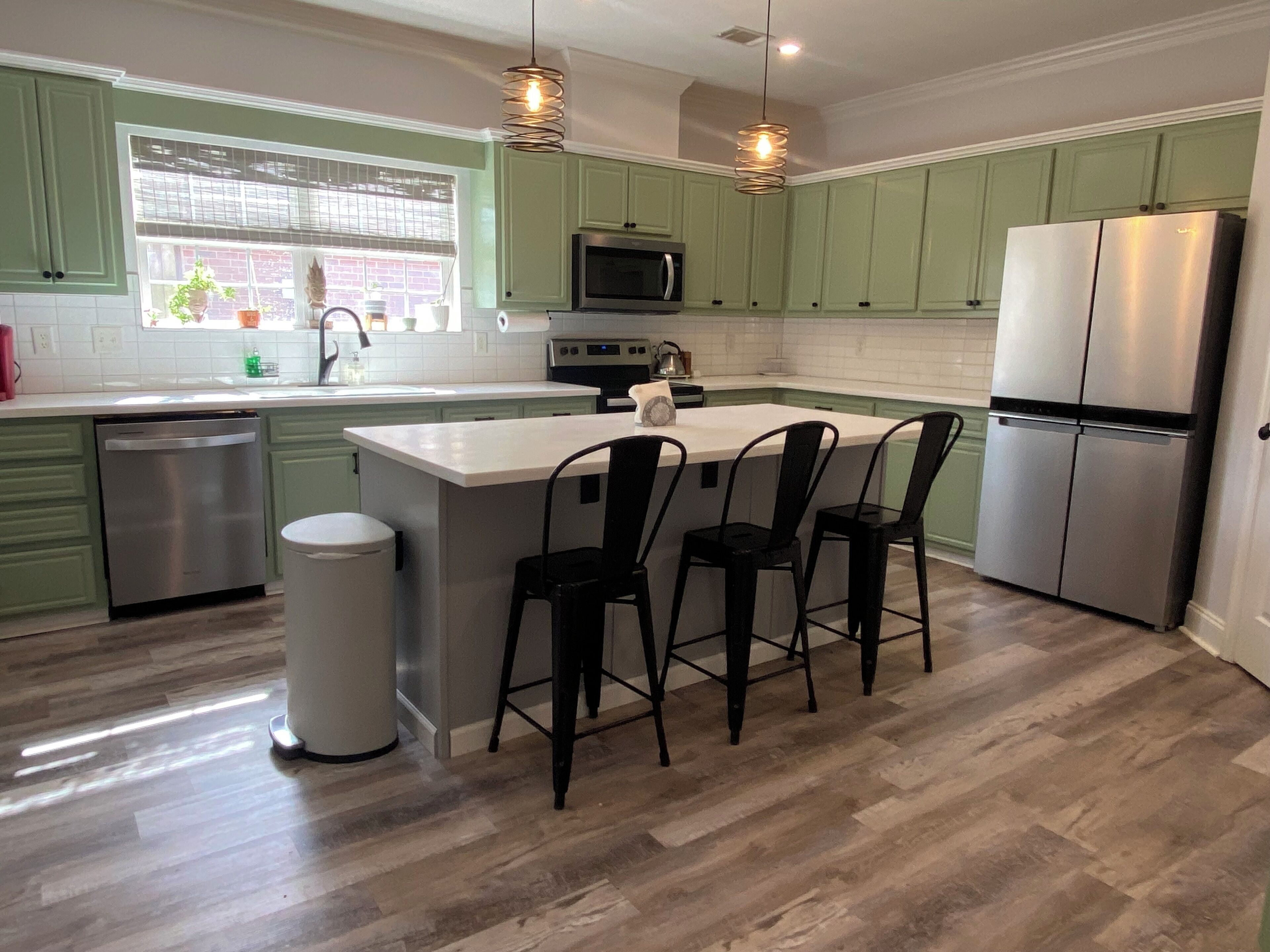 A modern kitchen with green cabinets, a stainless steel refrigerator, a microwave, and a sink. There are three black bar stools around the island.