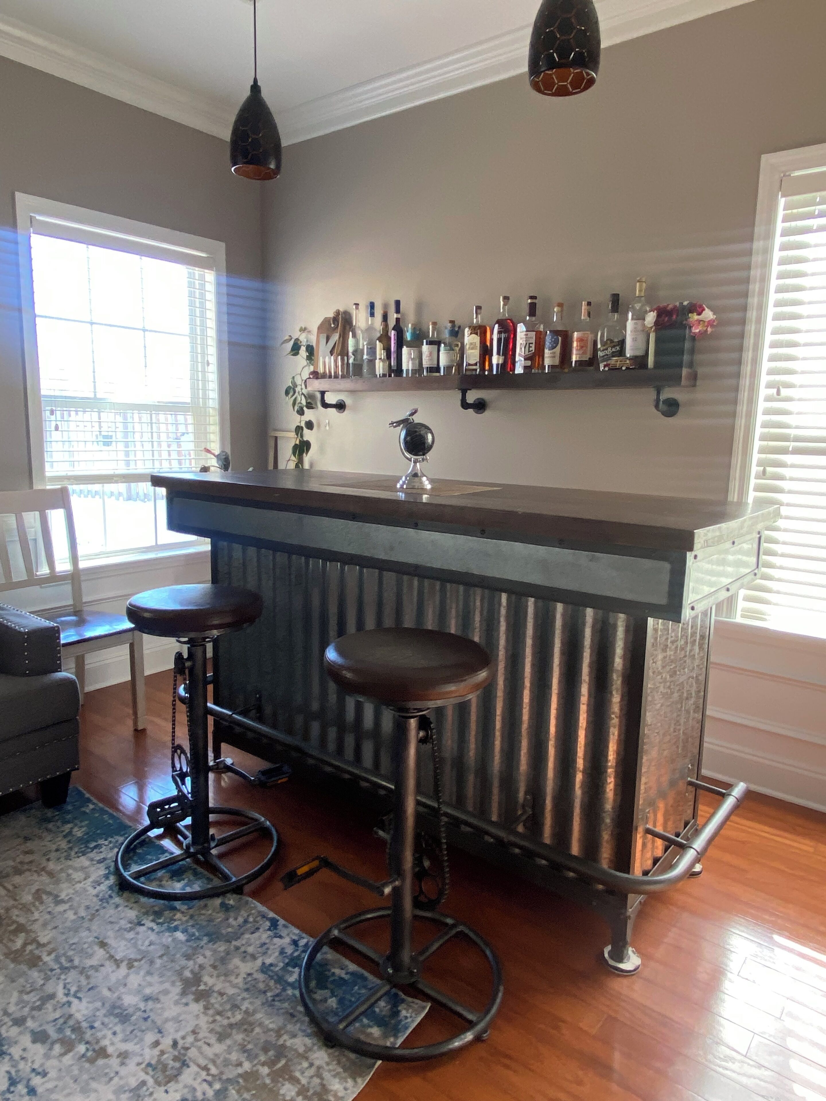 Un coin bar avec un comptoir en métal, deux tabourets, un luminaire suspendu et une étagère remplie de bouteilles.