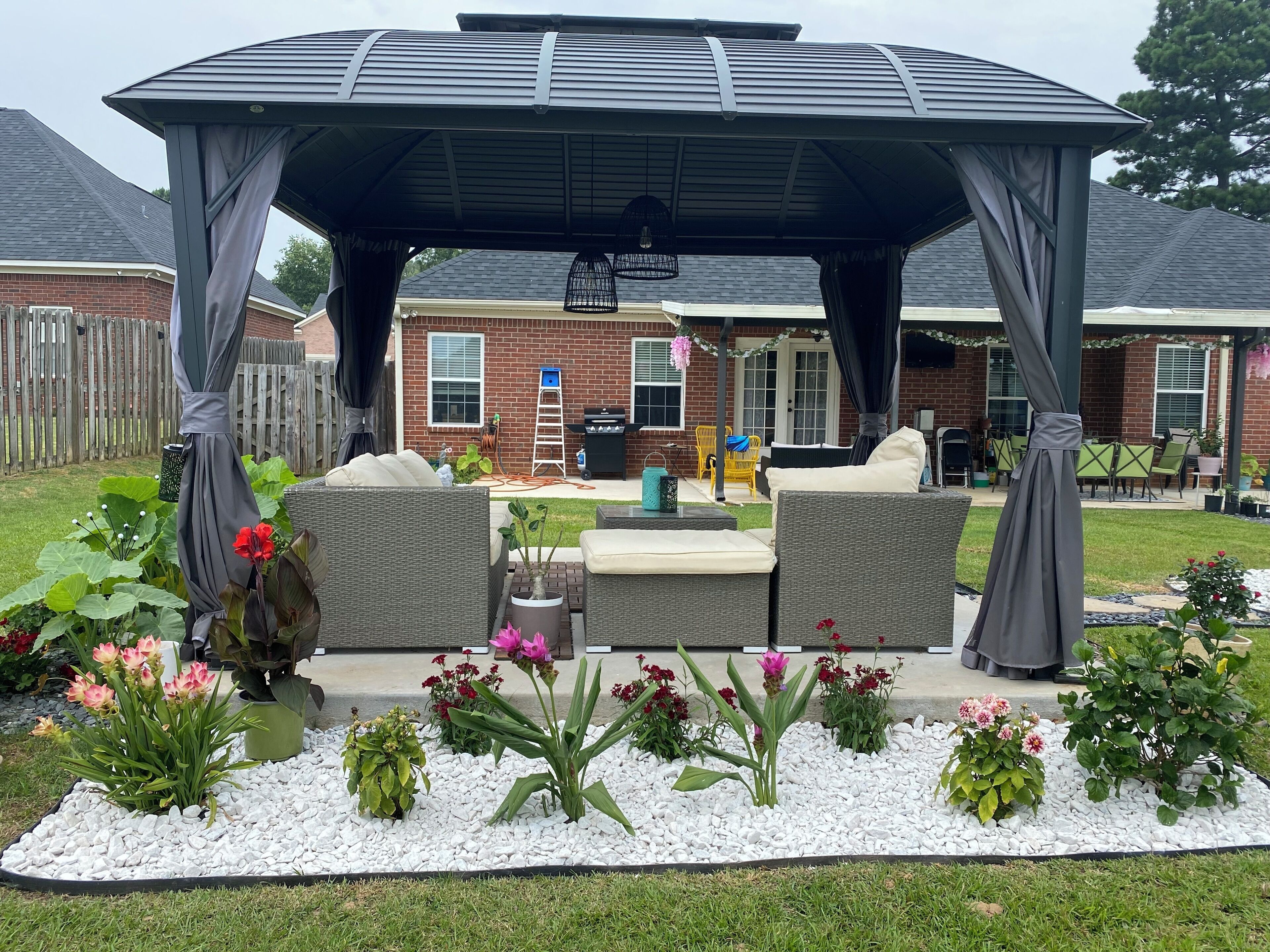Un patio coperto con arredi da esterno, una pergola e un giardino con diverse piante e fiori.
