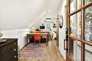 Une pièce compacte et bien éclairée, comprenant un bureau, une chaise et un lit, et agrémentée d’une grande fenêtre et d’un miroir décoratif.
