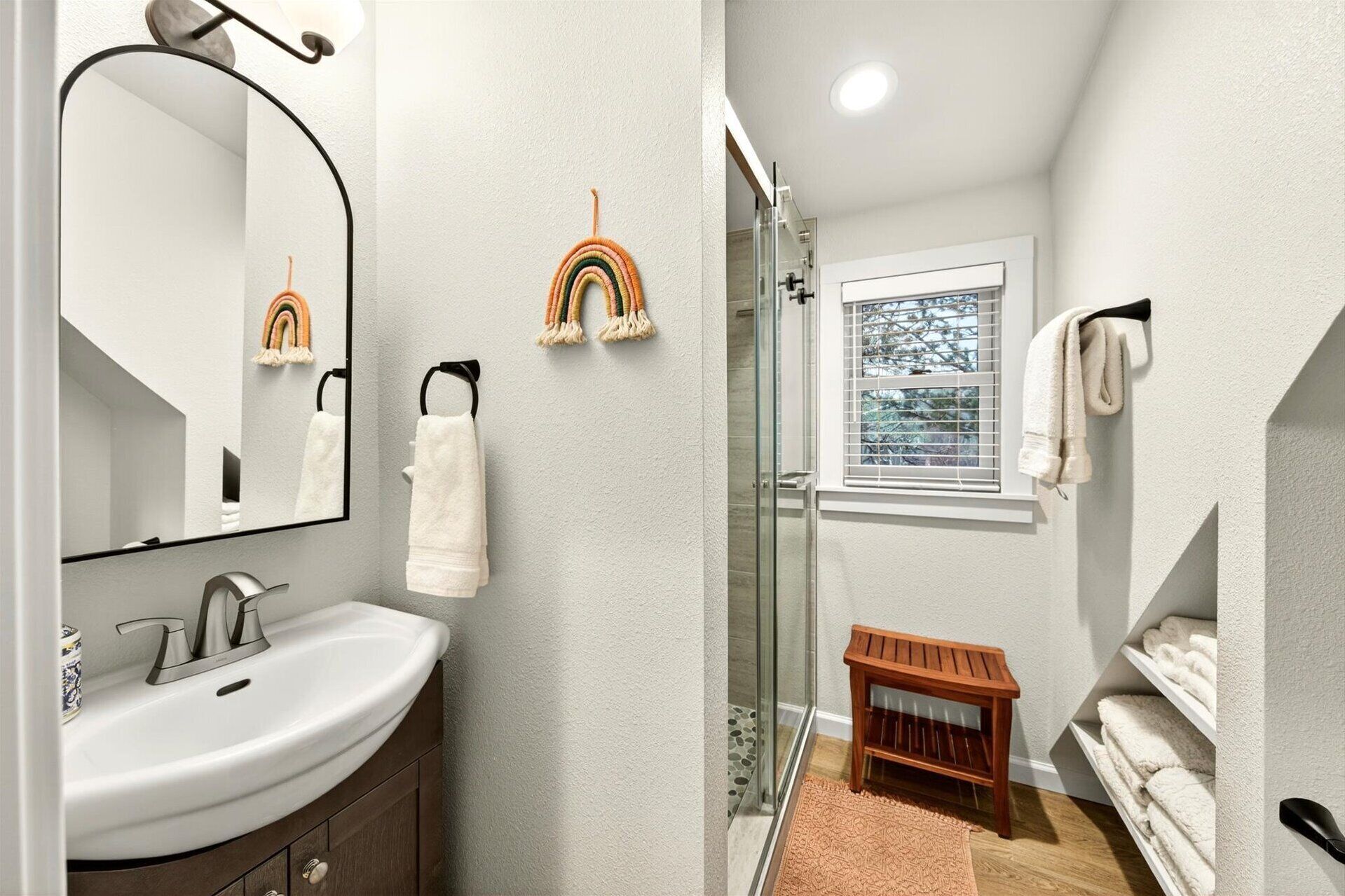 A bathroom with a white sink, a mirror, a glass shower enclosure, and a wooden bench.