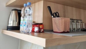 A kitchen shelf with a kettle, water bottles, a box of tea, a basket with utensils, and glass containers. - Mashley Room at Serpong Garden Apartment (Cisauk)
