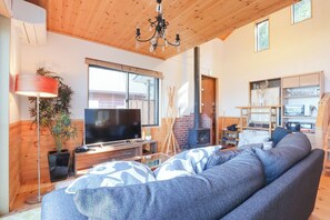 A cozy living room with a large sofa, a flat-screen TV, a wooden ceiling, and a fireplace. - Izu-atami Ocean View House (Atami)