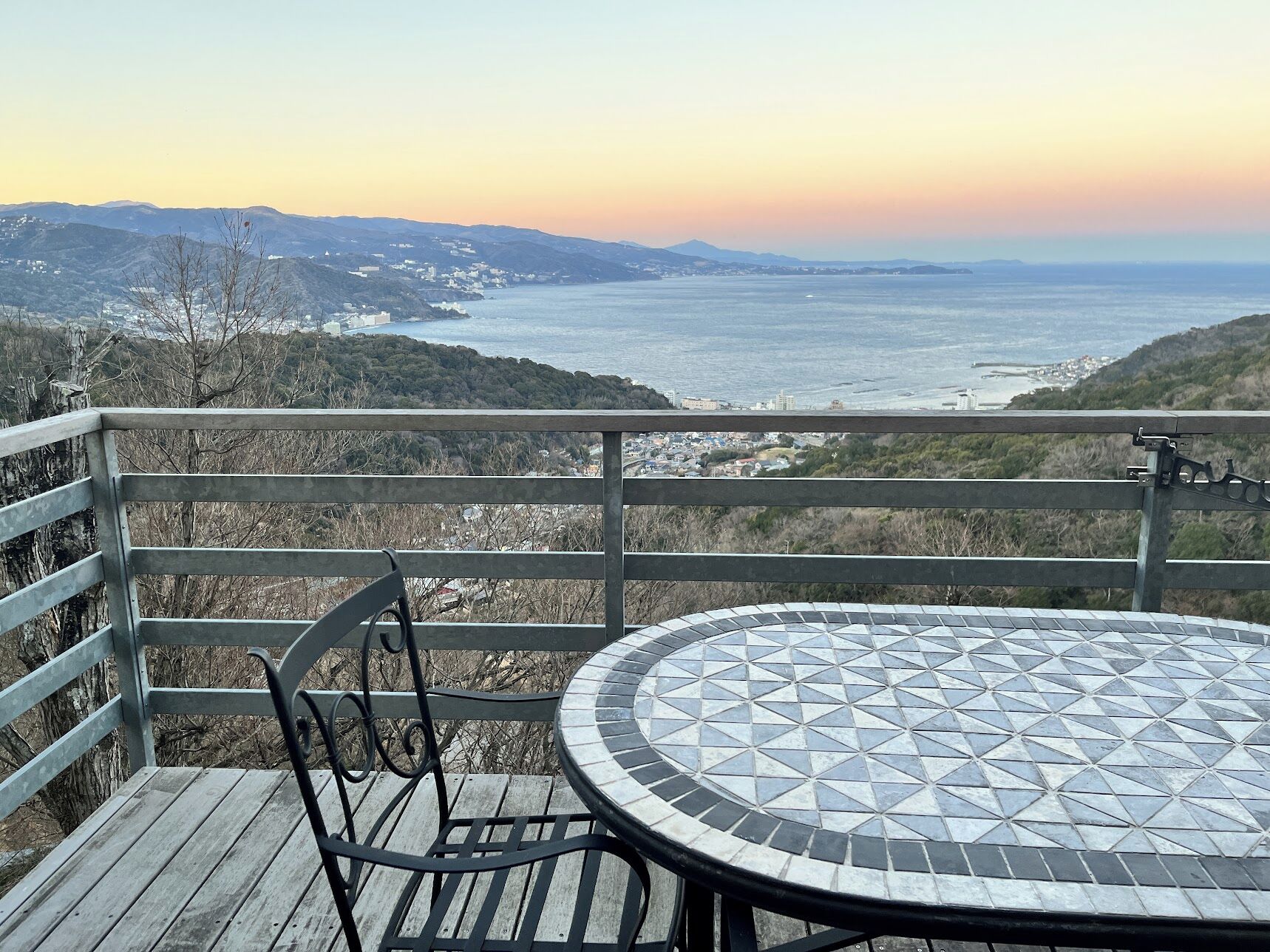 A wooden deck with a round table and chairs, overlooking a coastal landscape with hills and a view of the sea.