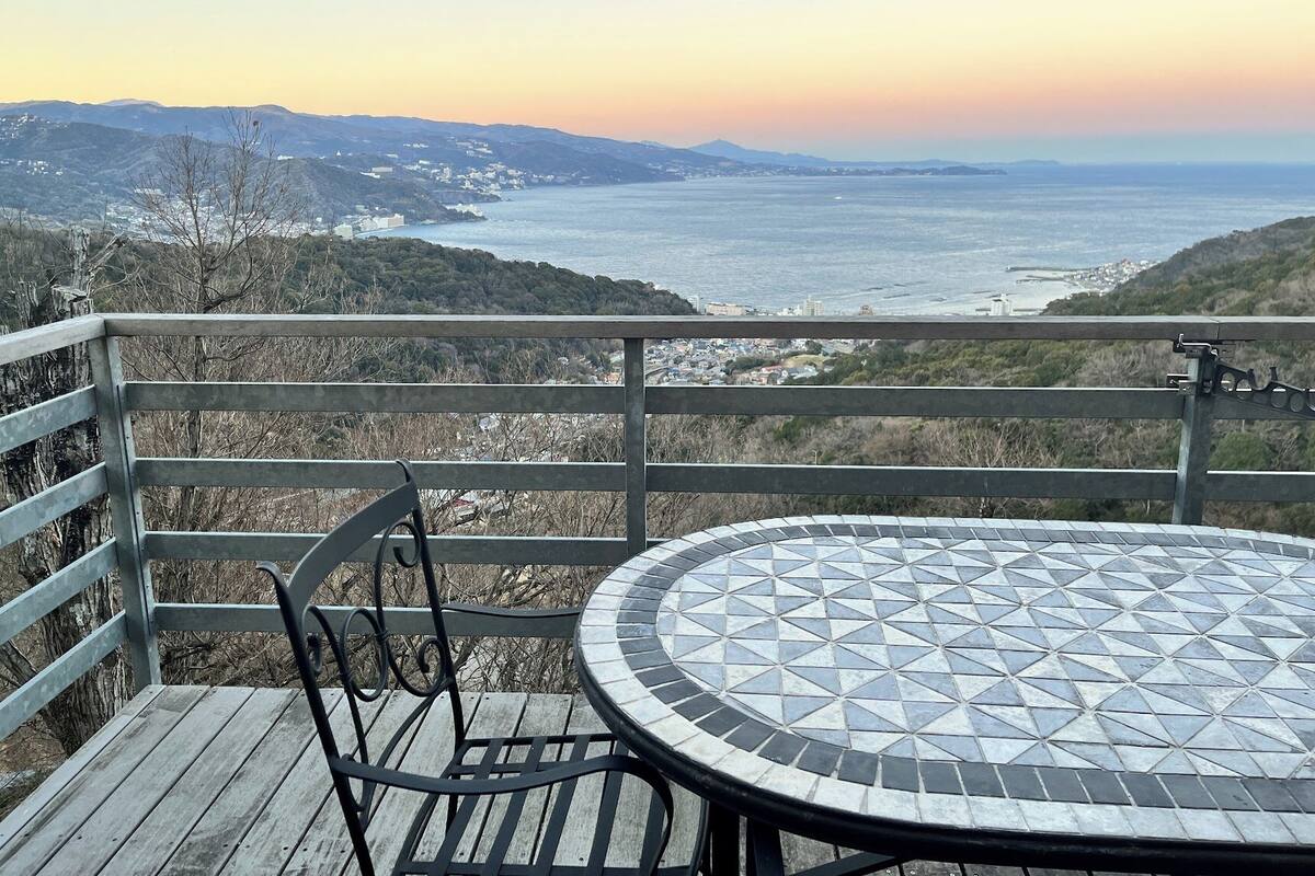 Une terrasse en bois avec une table ronde et des chaises, offrant une vue sur un paysage côtier vallonné et sur la mer.