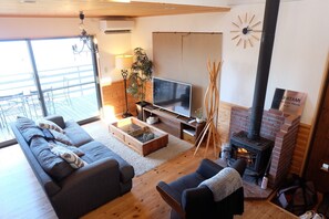 A modern living room with a sofa, TV, fireplace, and a balcony with outdoor furniture. - Izu-atami Ocean View House (Atami)