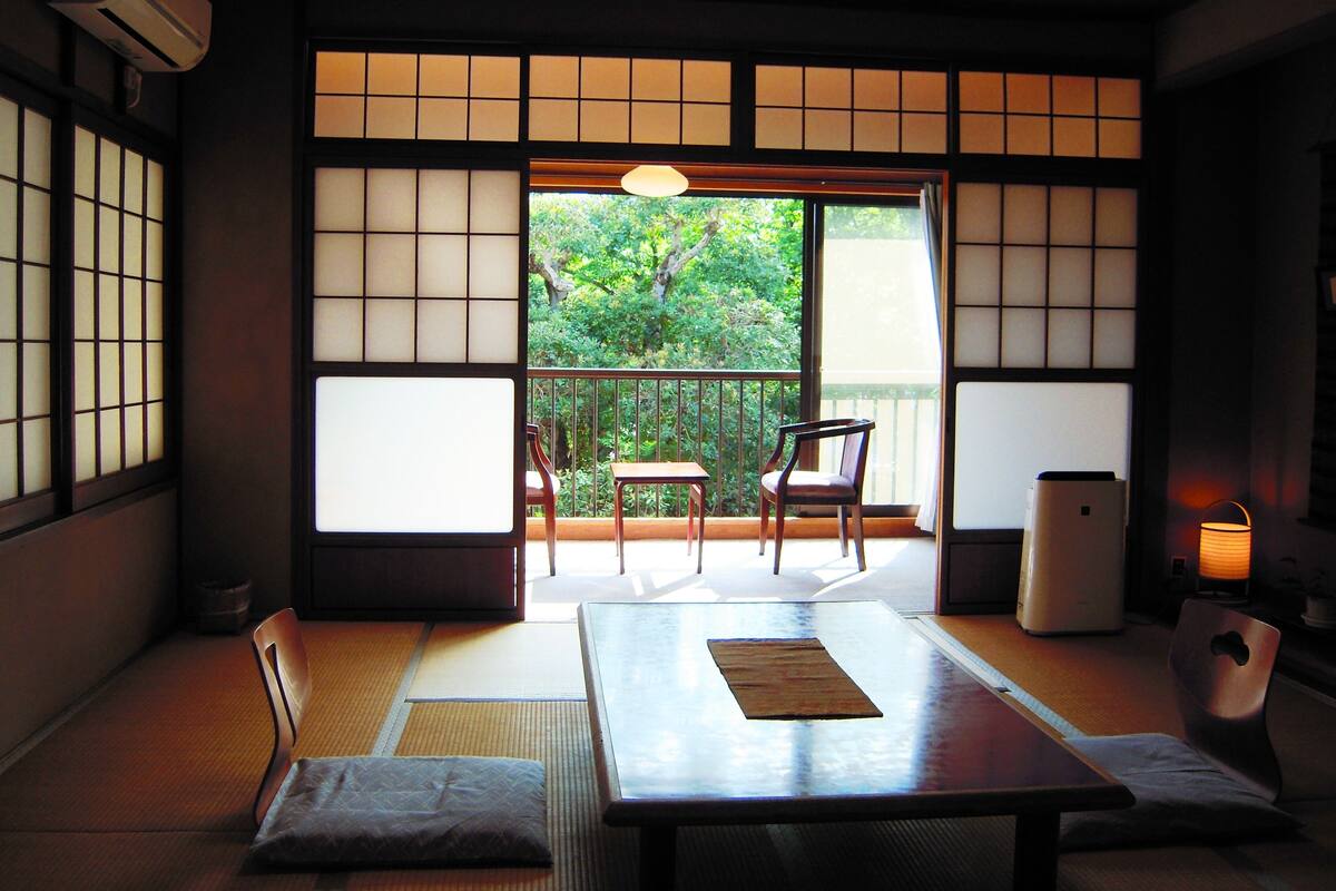 A traditional Japanese room with tatami flooring, a sliding door, a table, chairs, and a view of greenery outside.
