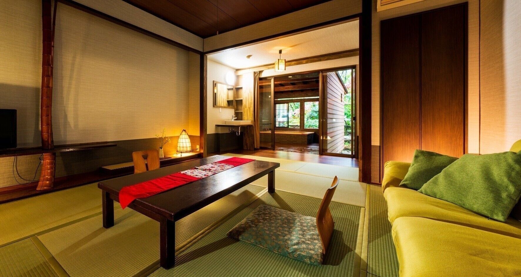 A traditional Japanese room with tatami flooring, a low table with a red runner, a yellow sofa, and a sliding door leading to a garden.