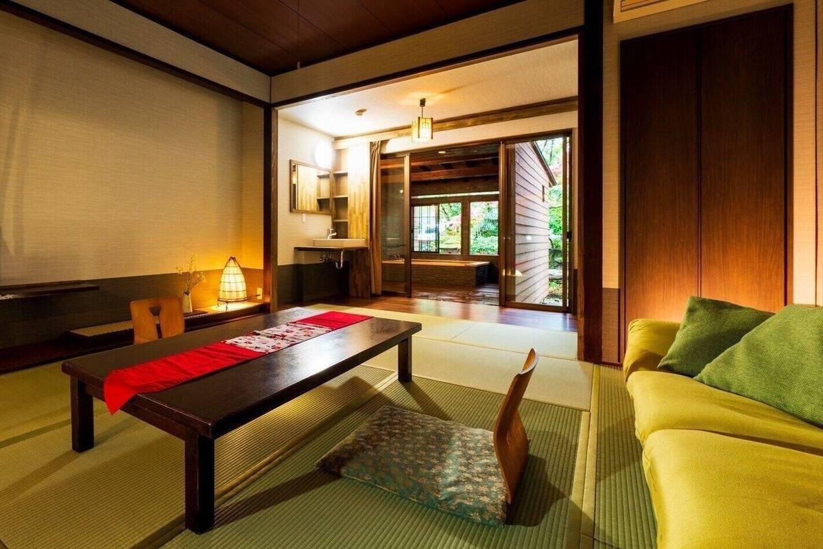 A traditional Japanese room with tatami flooring, a low table with a red runner, a yellow sofa, and a sliding door leading to a garden.