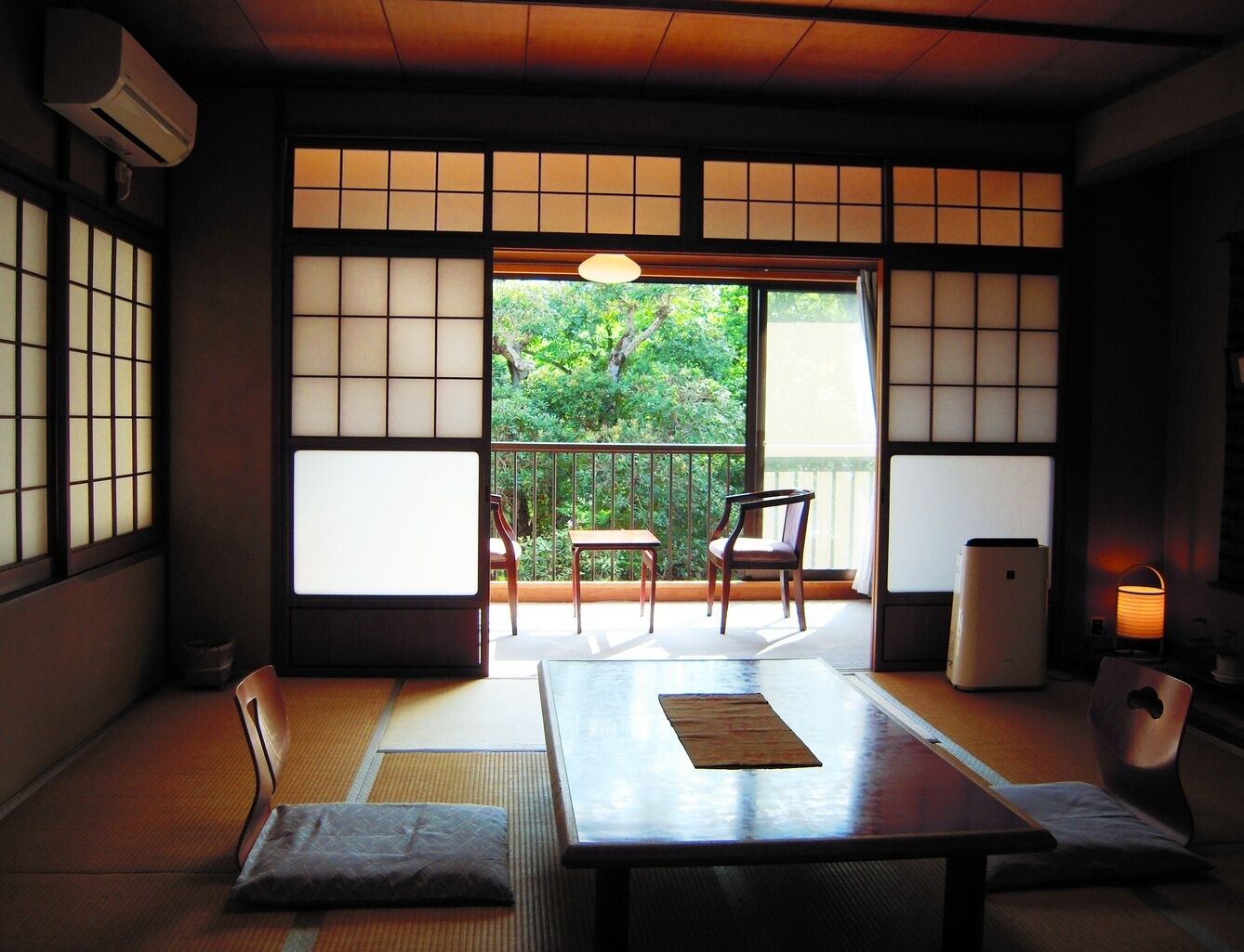 A traditional Japanese room with tatami flooring, sliding doors, a table with cushions, a chair, and a view of greenery outside.