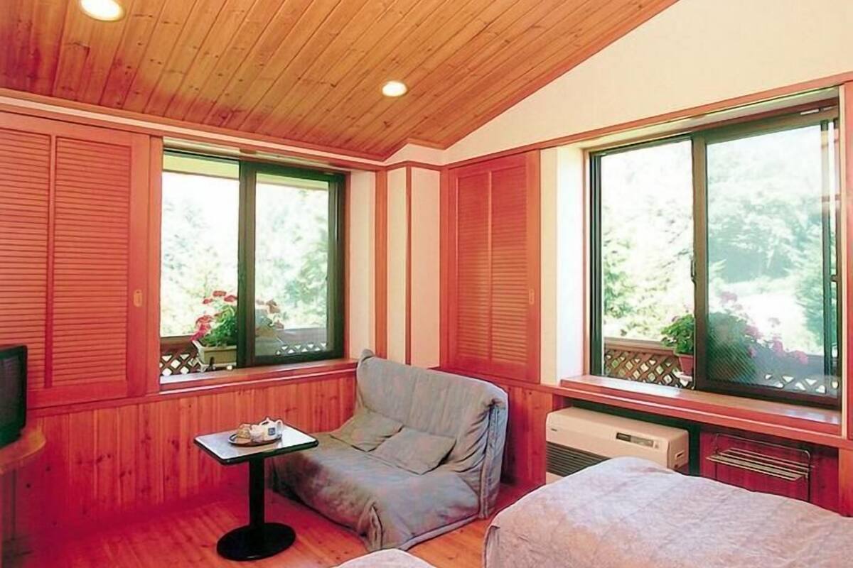 Um quarto aconchegante com paredes de madeira, um banco estofado, uma mesa pequena e janelas amplas com vista para o verde.