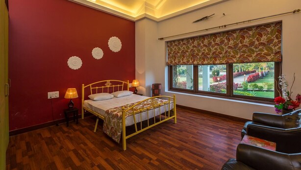 A bedroom with a wooden floor, a bed with a yellow frame, a window with a patterned curtain, and a view of a garden. - Amã£ Stays & Trails Nine Palms, Alibag (Alibag)