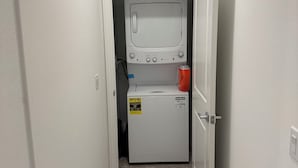 Une buanderie équipée d’un lave-linge et d’un sèche-linge à chargement frontal, d’une bouche d’aération au-dessus du sèche-linge et d’une porte donnant sur une autre pièce.