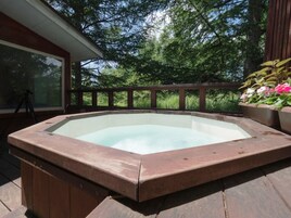 Un jacuzzi en una terraza de madera con barandilla y una ventana con vista a un área boscosa.