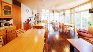 A spacious dining area with wooden tables and chairs, a kitchen area with wooden cabinets, and large windows providing natural light. - Guesthouse Curry Village (Kiryu)