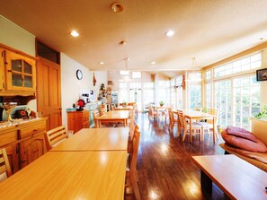A spacious dining area with wooden tables and chairs, a kitchen area with wooden cabinets, and large windows providing natural light. - Guesthouse Curry Village (Kiryu)