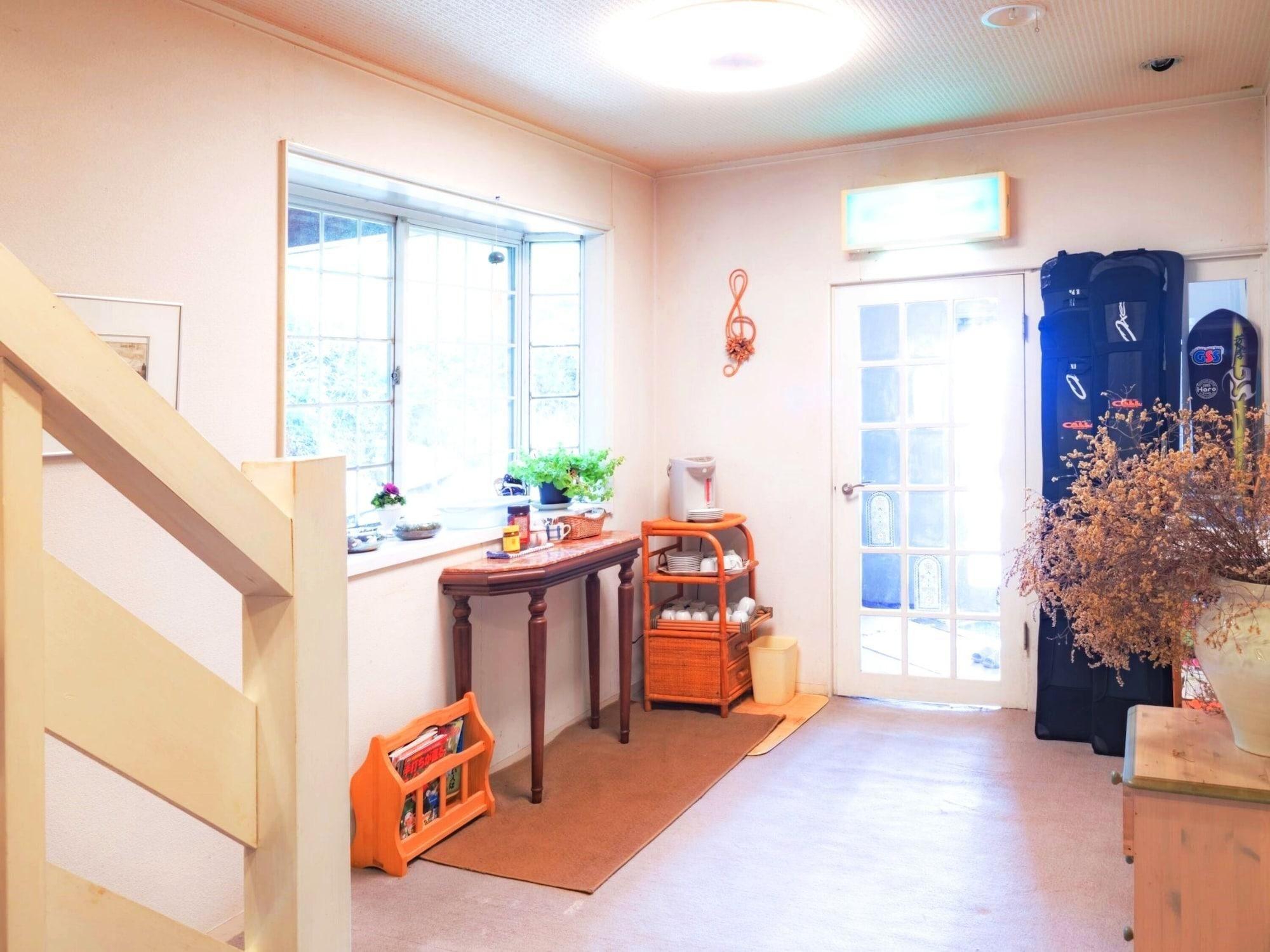 A bright room with a wooden staircase, a window with plants, a small table with a basket, and a door with a blue bag hanging on it.