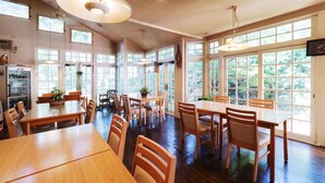 A spacious dining area with multiple wooden tables and chairs, large windows offering views of greenery, and ceiling lights. - Guesthouse Curry Village (Kiryu)