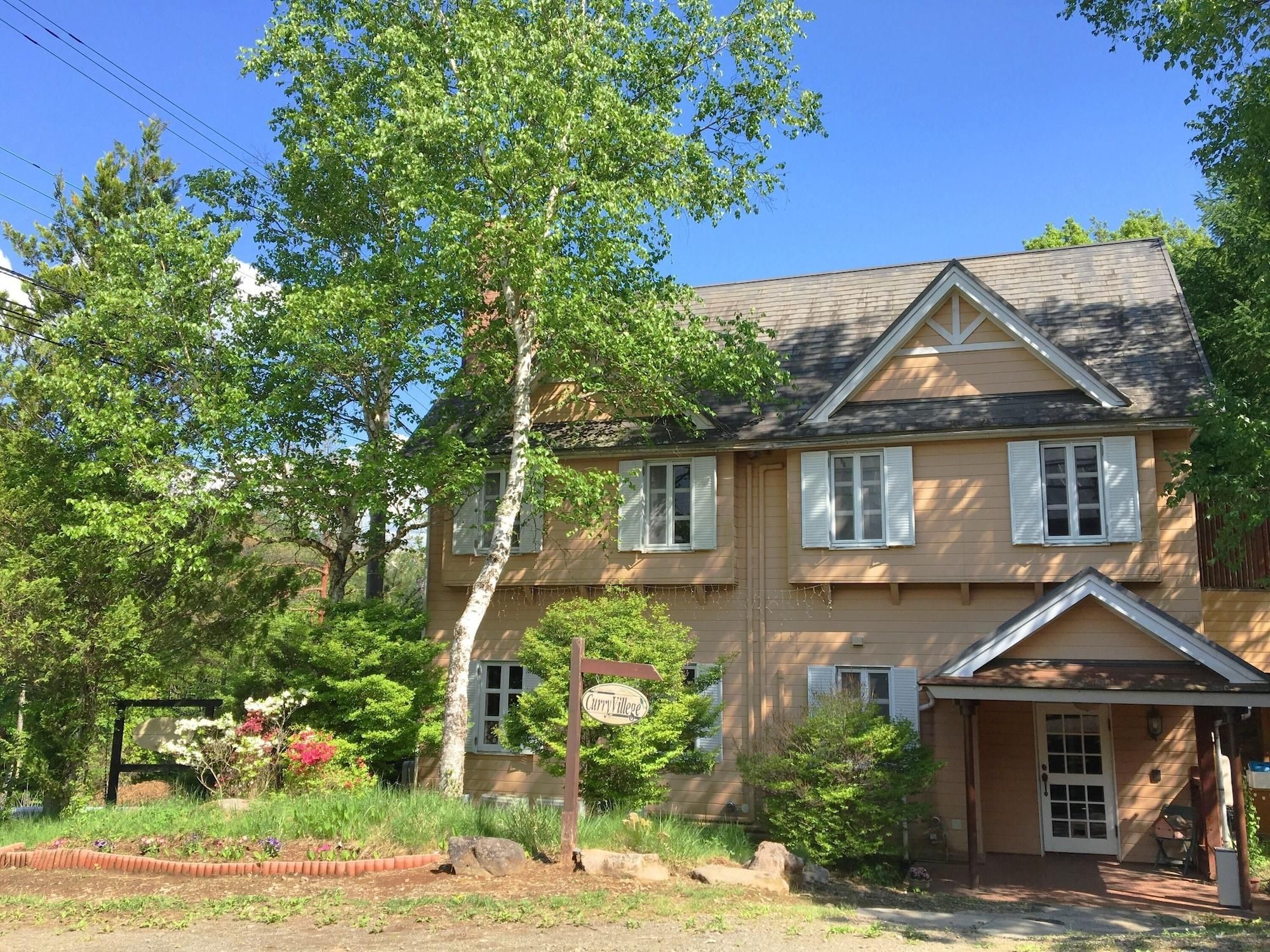 Une maison à deux étages avec un porche à l’avant, une enseigne indiquant « Curry Village », et un jardin bien entretenu où poussent différentes plantes et fleurs.