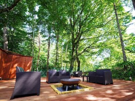 Un patio al aire libre con mobiliario de mimbre, un pozo central para fuego y una terraza de madera rodeada de árboles.