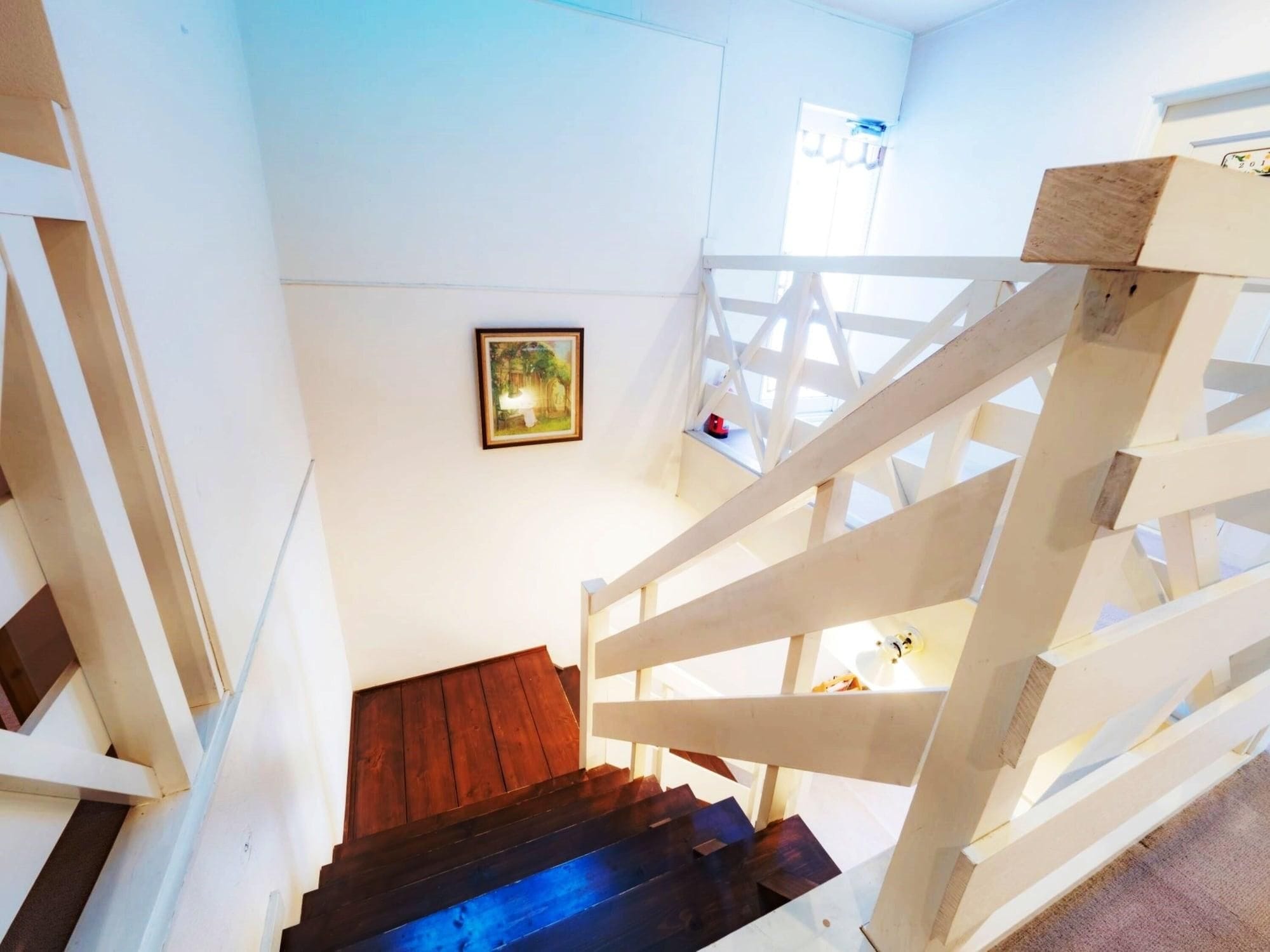 Un escalier en bois, doté d’une main courante en bois foncé et de balustrades blanches, mène à un palier où se trouvent un tableau encadré et une petite fenêtre.