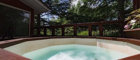 A hot tub surrounded by a wooden deck with a glass enclosure, set against a backdrop of tall trees.