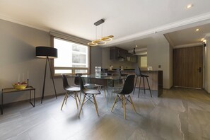 A modern dining area with a glass table and black chairs, a black floor lamp, and a view of the kitchen and balcony. - Capitalia Living Apartments J Verne (Mexico City)