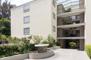 A multi-story building with a courtyard featuring a central fountain, surrounded by greenery and a glass-roofed upper level with balconies. - Capitalia Living Apartments J Verne (Mexico City)