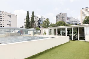 A rooftop terrace with a glass enclosure, a small lawn, and a building with a glass facade. - Capitalia Living Apartments J Verne (Mexico City)