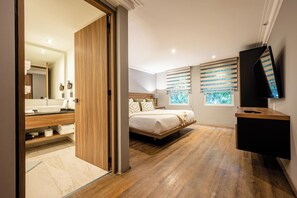 A modern bedroom with a wooden floor, a bed with a wooden headboard, a bedside table, a TV, and a window with blinds. - Capitalia Living Apartments J Verne (Mexico City)