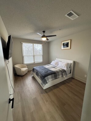 A bedroom with a bed, a ceiling fan, a window with blinds, a chair, and a wall-mounted TV.