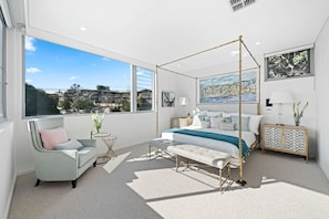 A modern bedroom with a four-poster bed, a sitting area with a chair and a small table, and a view of the cityscape through large windows. - Fully Furnished house 89 day option (Vaucluse)