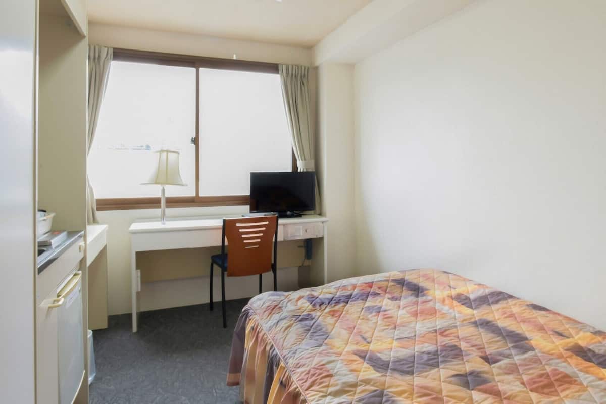 A small hotel room with a bed, a desk with a computer, a chair, and a window with curtains.