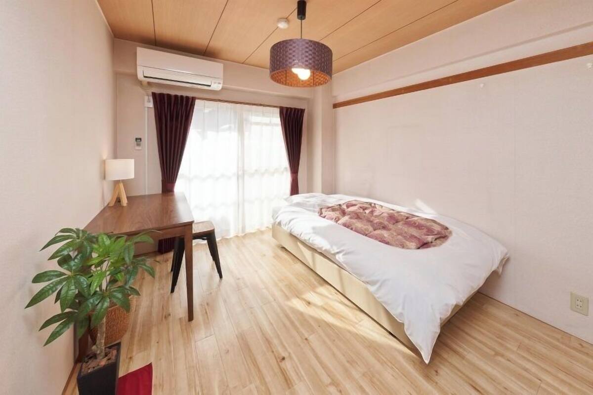 Ein Schlafzimmer mit Bett, Schreibtisch, Lampe, einem Topf mit Pflanze und einer Klimaanlage.