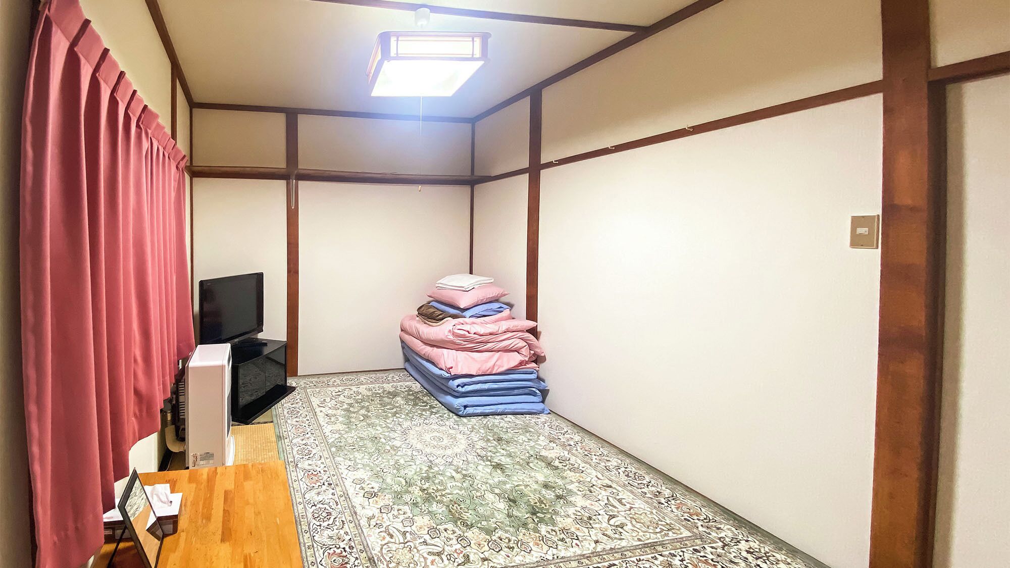 A room with a television, a stack of folded bedding, a patterned rug, and a wooden-framed ceiling.