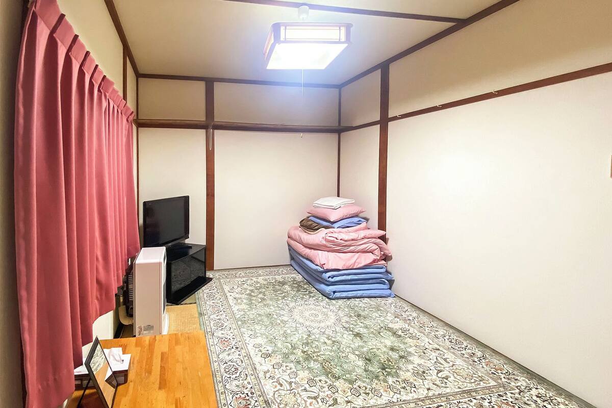 Een kamer met een televisie, een stapel opgevouwen beddengoed, een kamerkleed met patroon en een plafond met houten balken.