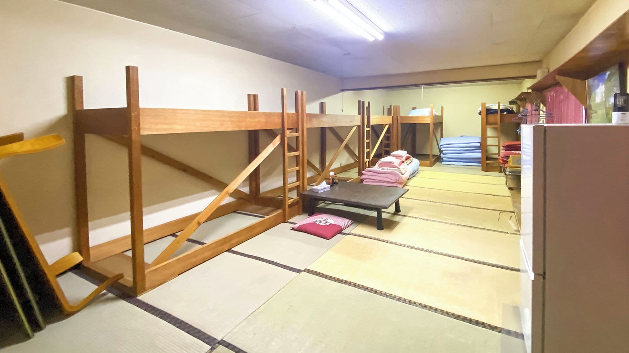 A room with tatami flooring, wooden bunk beds, a small table with a pillow, and a refrigerator.